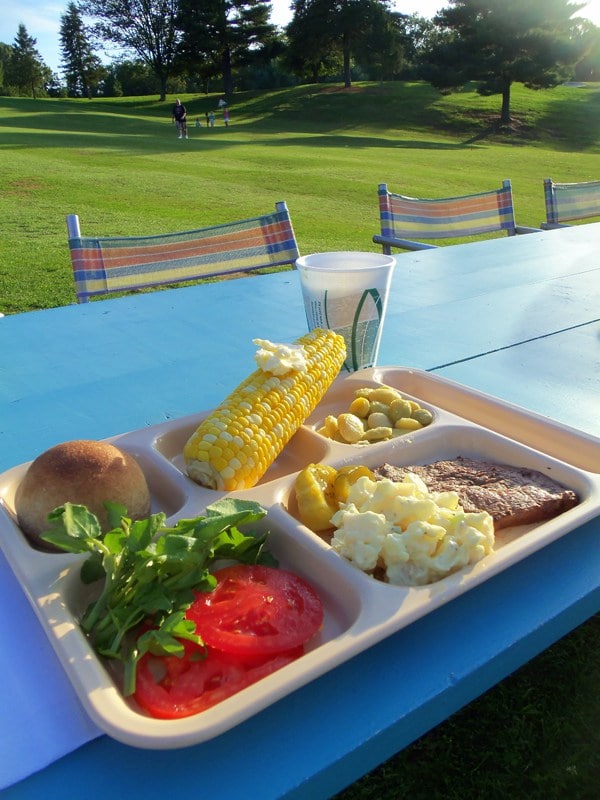 tray of food with buttered corn on the cob, tomatoes, lettuce, a roll, potato salad and steak